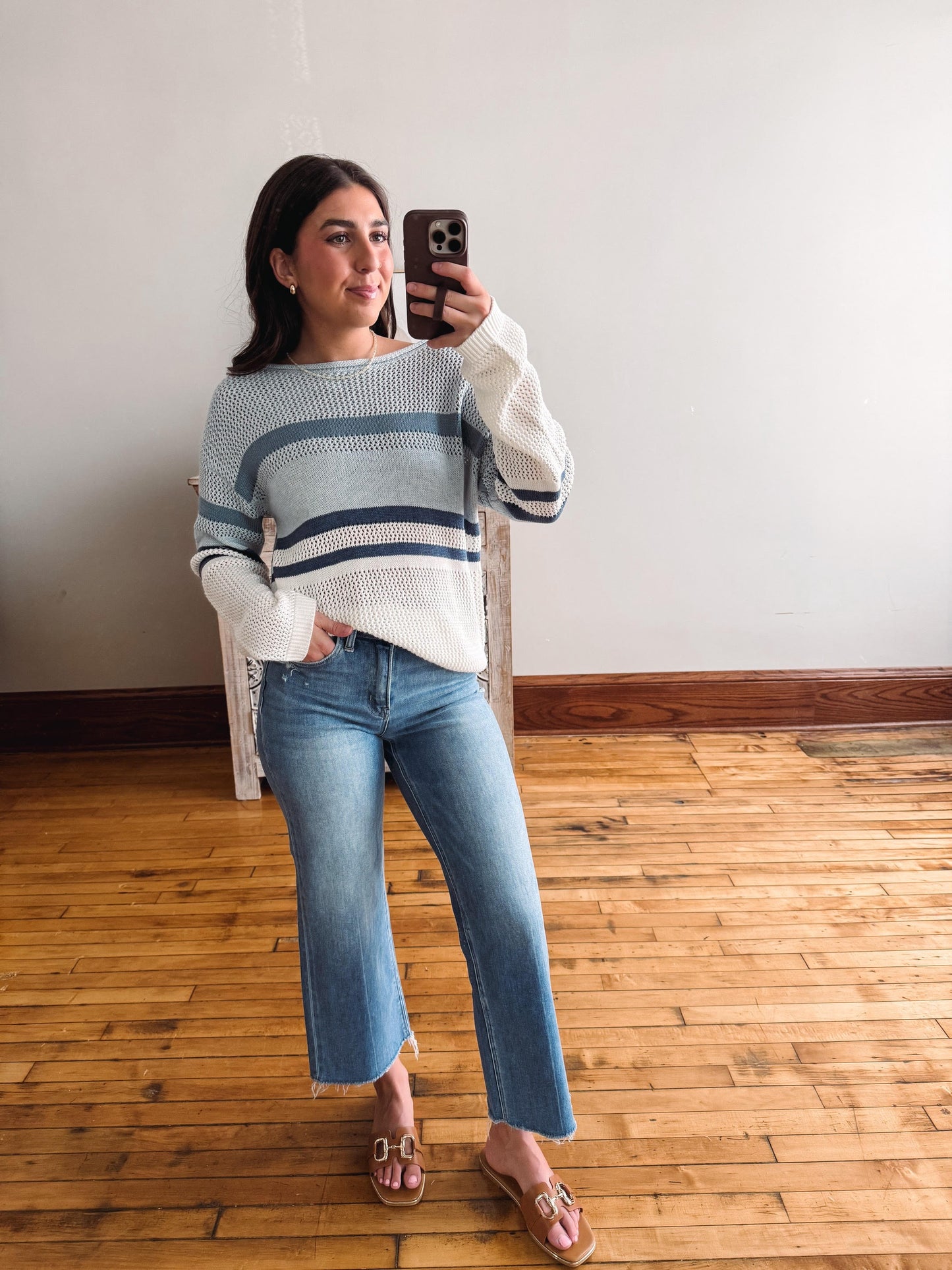 Woman taking a mirror selfie wearing a striped sweater and jeans in a room with wooden flooring.