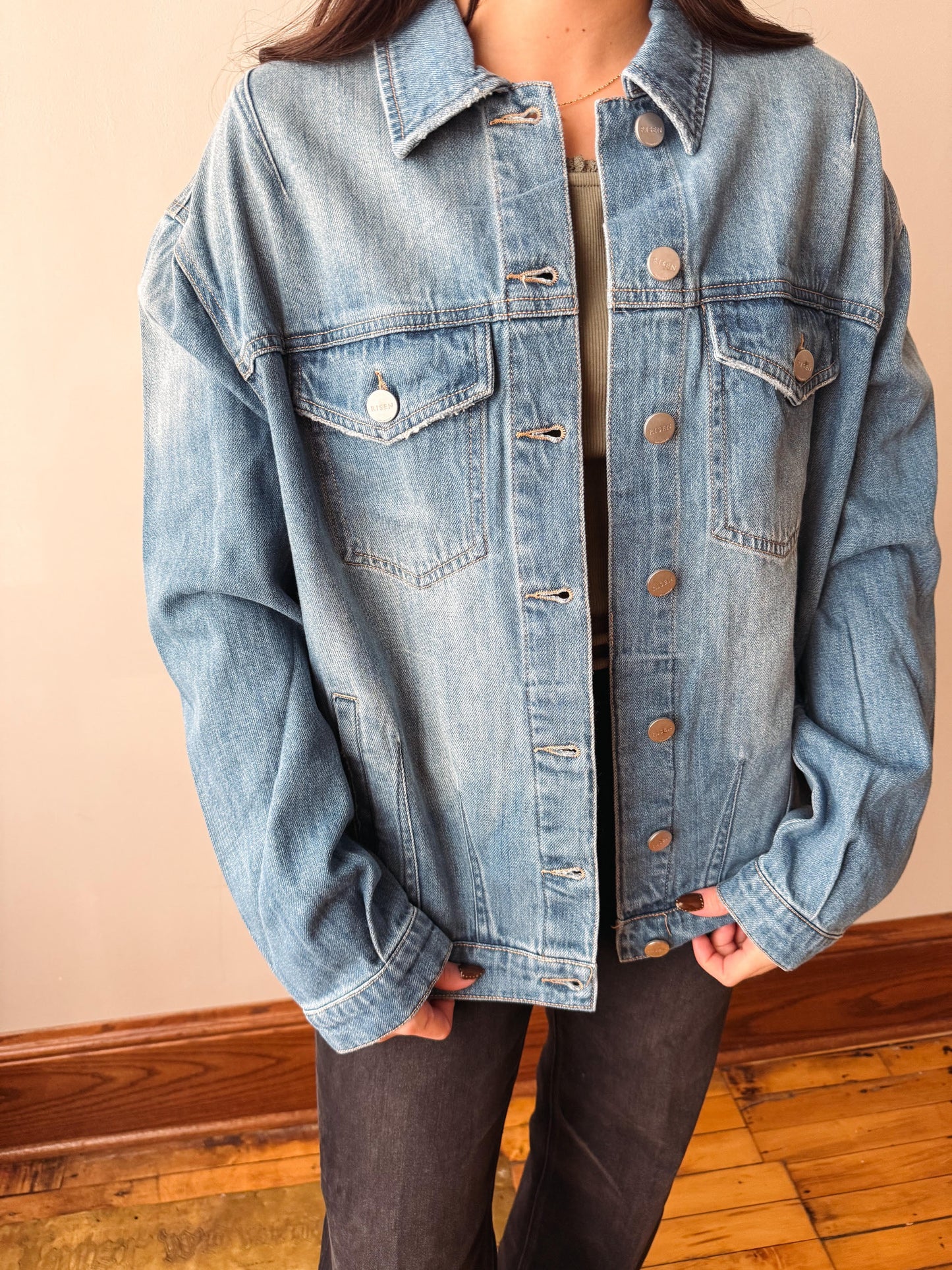 A person wearing an oversized light-wash blue denim jacket with front button closure, chest flap pockets, and contrast stitching, paired with dark pants, standing indoors against a beige wall and wood floor.