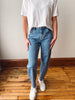 A person wearing light blue denim jeans with small white embroidered details, styled with a white t-shirt and white sneakers, standing on a wooden floor against a plain wall.