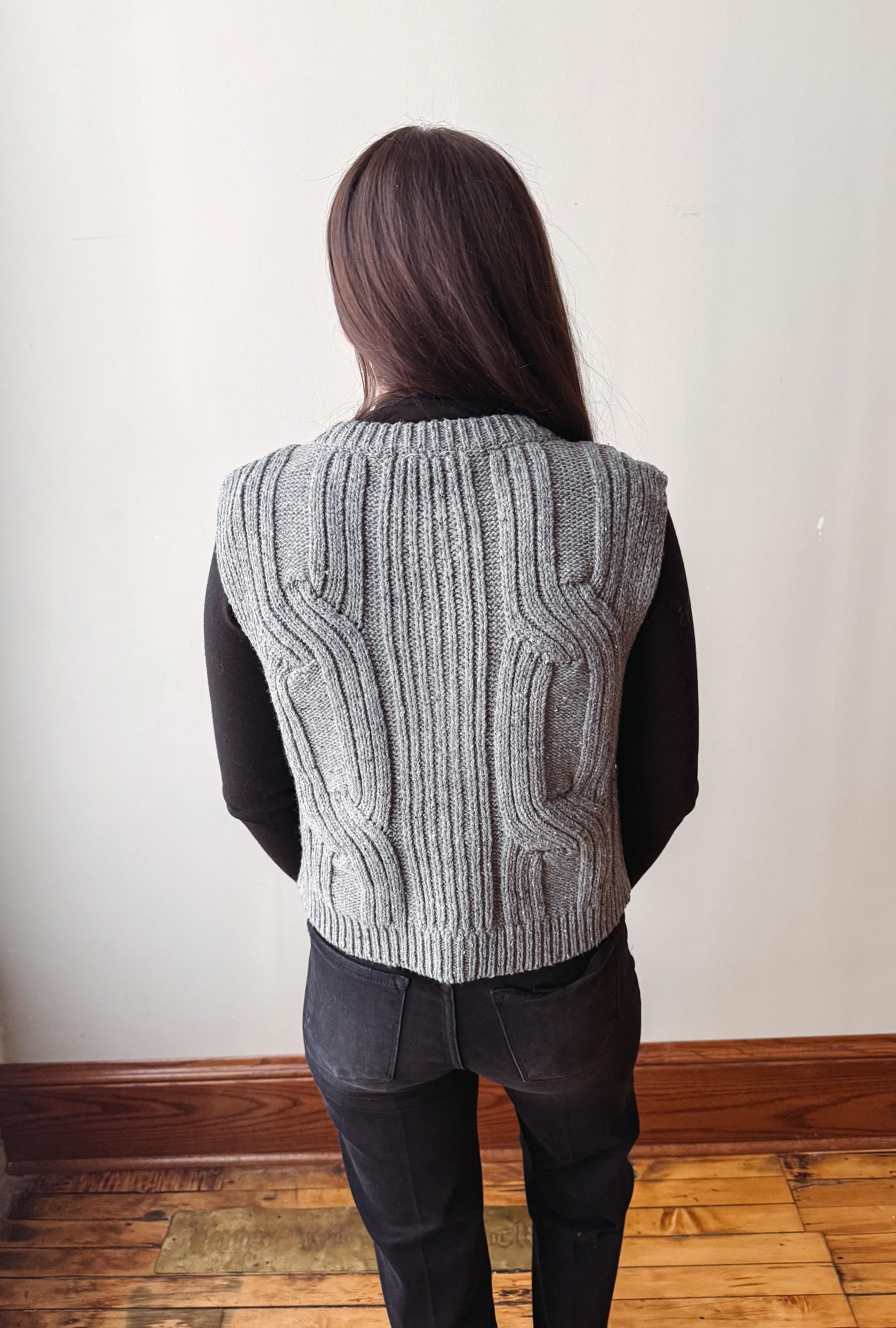 A person shown from behind wearing a gray cable-knit sweater vest over a black long-sleeve top and black jeans, standing in an indoor room with a light wall and wood floor.