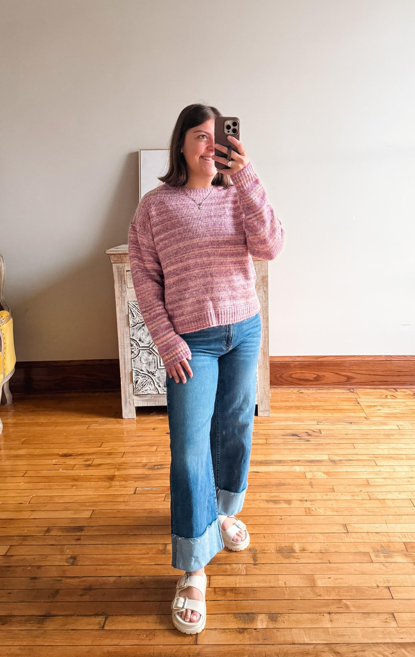 A woman takes a mirror selfie in a room with hardwood floors, wearing a pink and purple marled knit sweater, blue jeans, and white slide sandals.