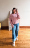 A woman takes a mirror selfie in a room with hardwood floors, wearing a pink and purple marled knit sweater, blue jeans, and white slide sandals.