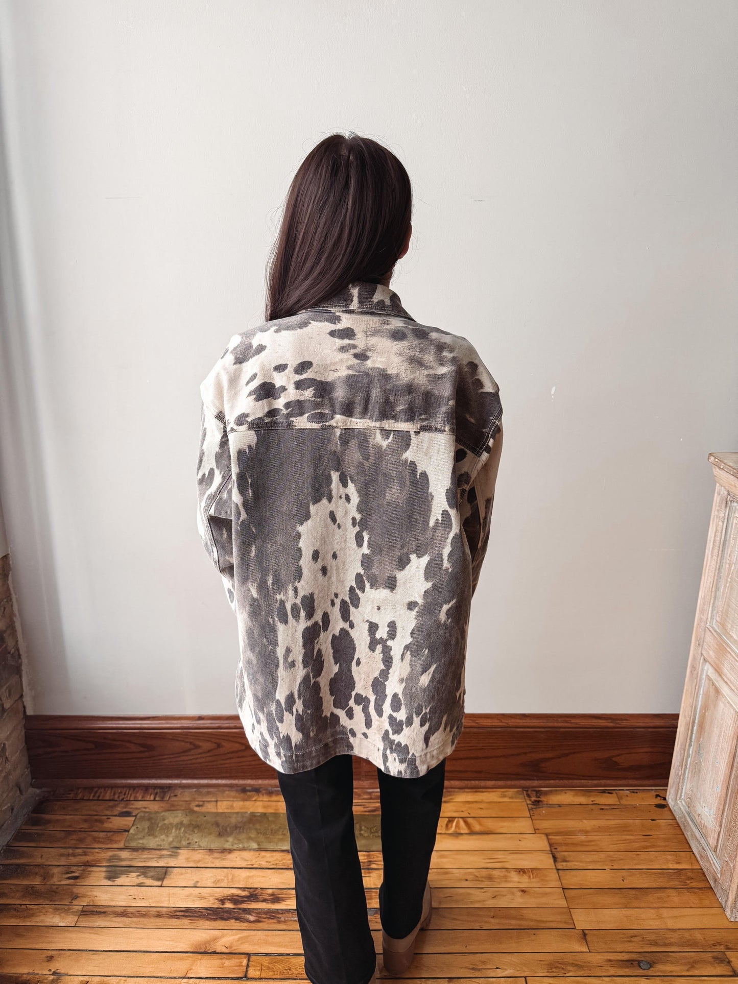 A person viewed from behind wearing a loose, oversized button-up shirt with a gray, black, and off-white tie-dye or mottled pattern, paired with black pants, standing on a wooden floor against a plain wall.