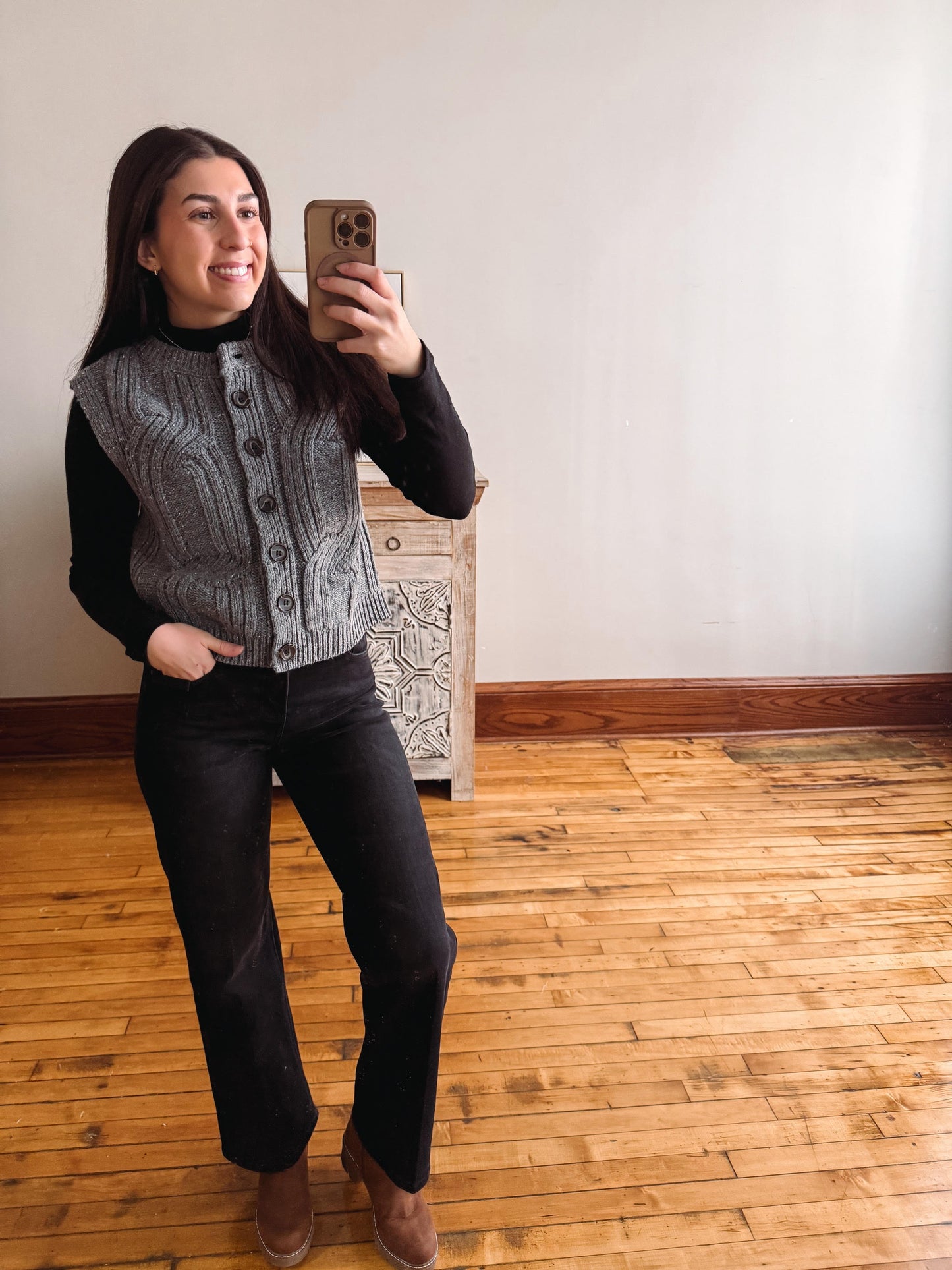 A woman takes a mirror selfie in a room with light wood floors and a white wall, wearing a gray cable-knit button-front vest over a black long-sleeve top and black pants.