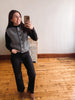 A woman takes a mirror selfie in a room with light wood floors and a white wall, wearing a gray cable-knit button-front vest over a black long-sleeve top and black pants.