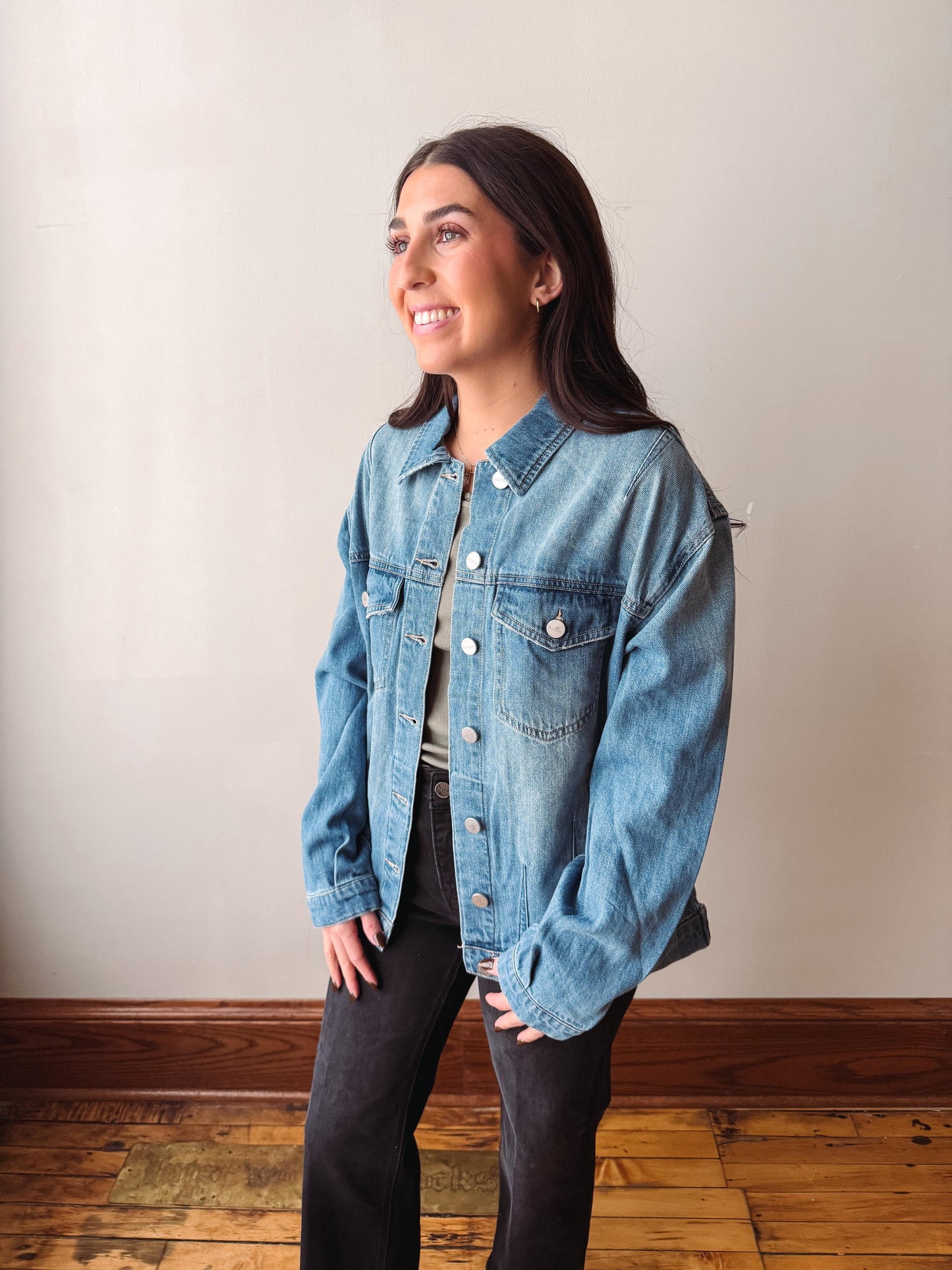 A woman wearing a light blue denim jacket over a white top and black pants, standing in a room with a beige wall and wooden floor.