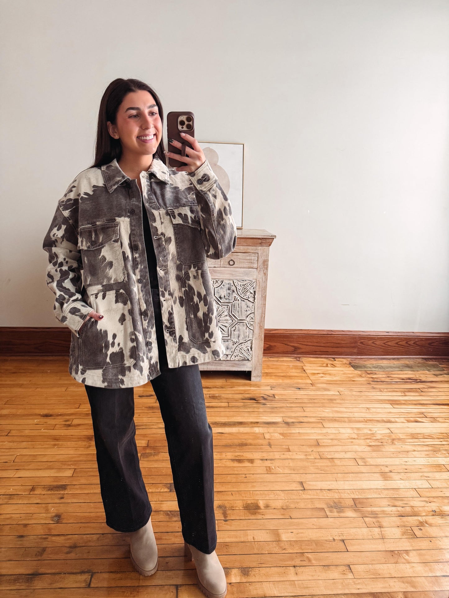 A woman takes a mirror selfie wearing a gray and white tie-dye patterned button-up shirt jacket, black wide-leg jeans, and beige ankle boots in a room with light wood floors and a white wall.