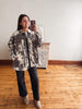 A woman takes a mirror selfie wearing a gray and white tie-dye patterned button-up shirt jacket, black wide-leg jeans, and beige ankle boots in a room with light wood floors and a white wall.