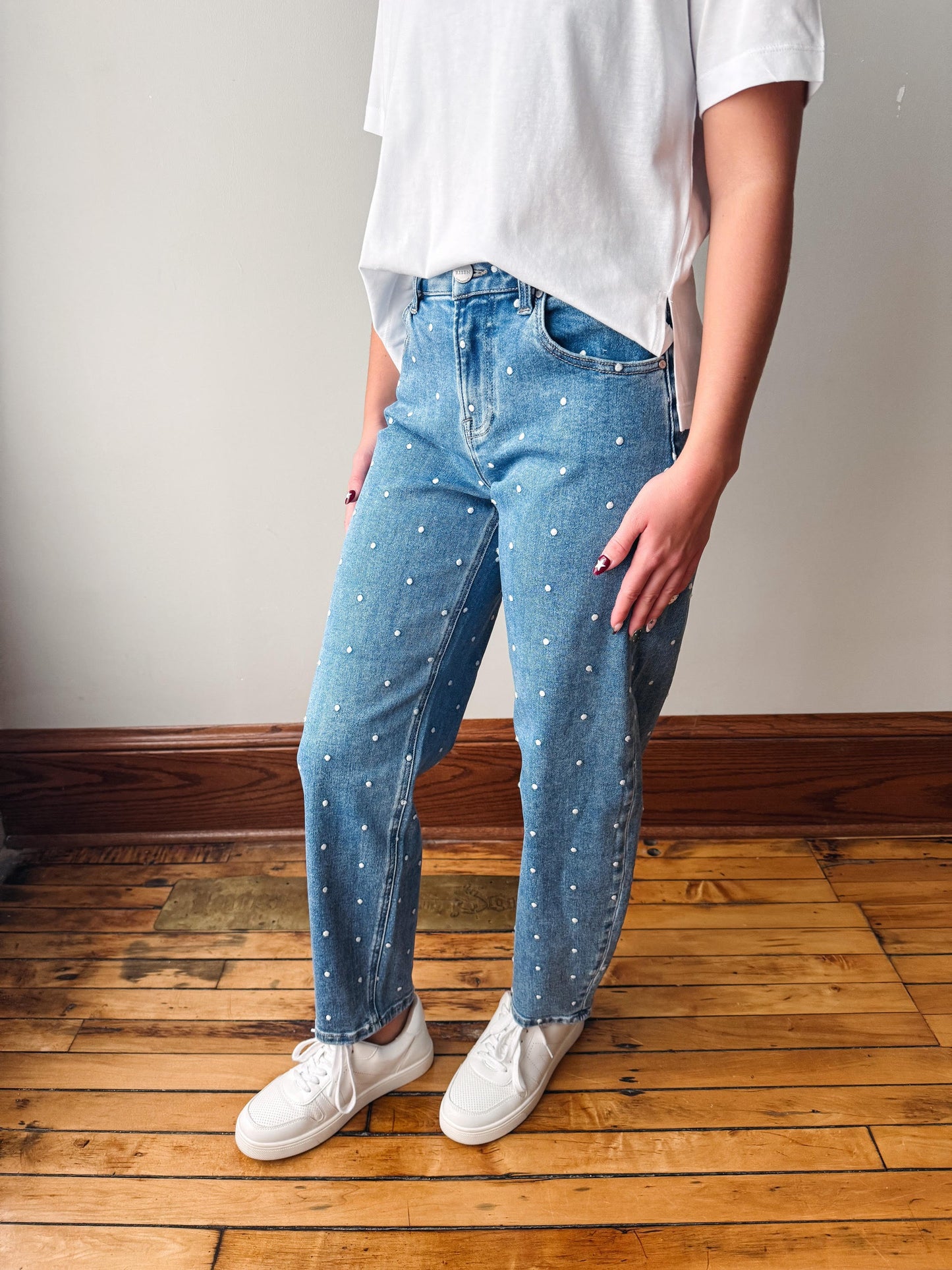 A person wearing light blue denim jeans with small white embroidered dots, styled with a white t-shirt and white sneakers, standing on a wooden floor against a plain wall.