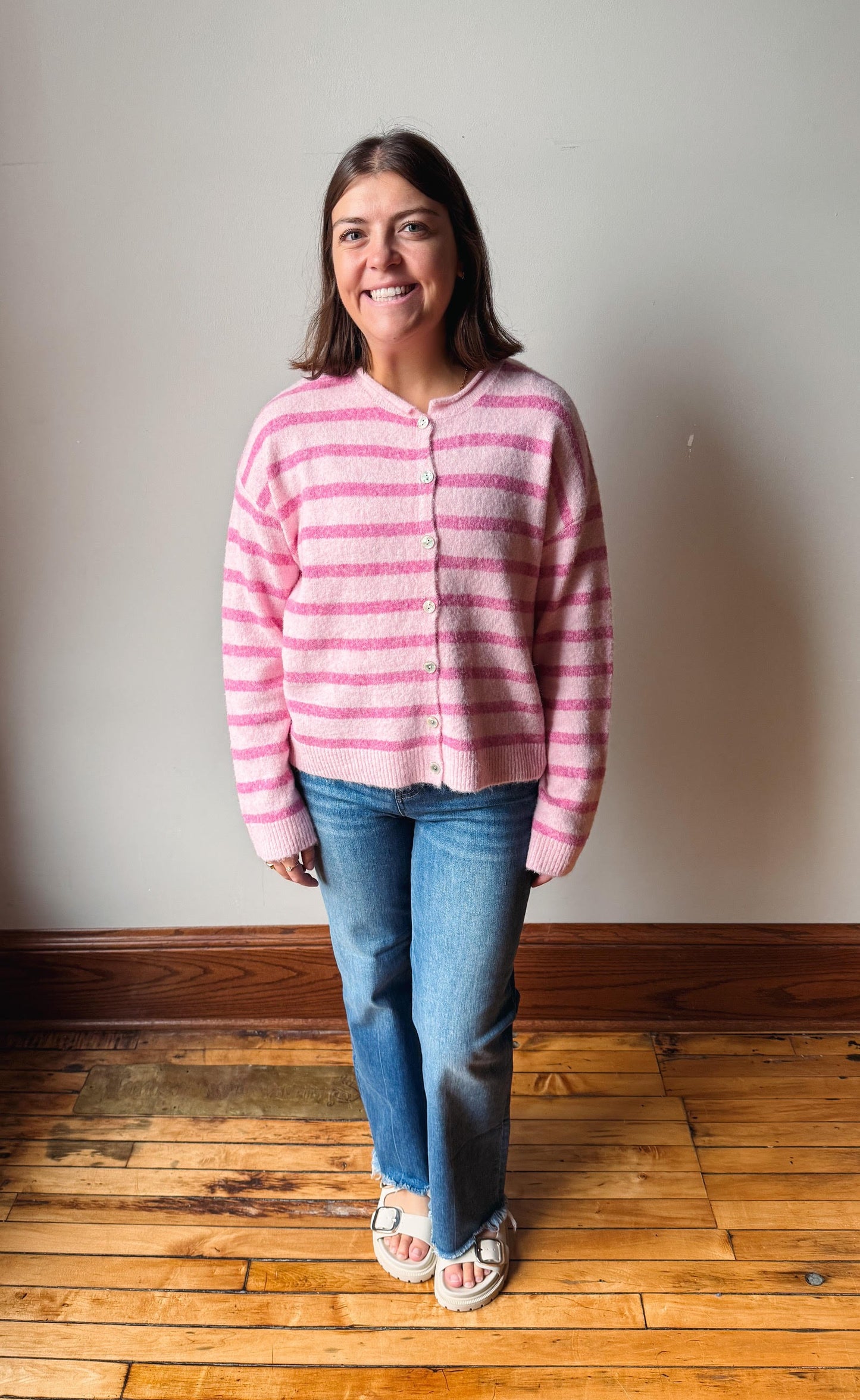 A woman standing in a room with a light gray wall and wooden floor, wearing a pink and white striped fuzzy cardigan, blue jeans, and white slide sandals.