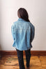 A person viewed from behind wearing an oversized light blue denim jacket and dark pants, standing on a wooden floor against a plain off-white wall.