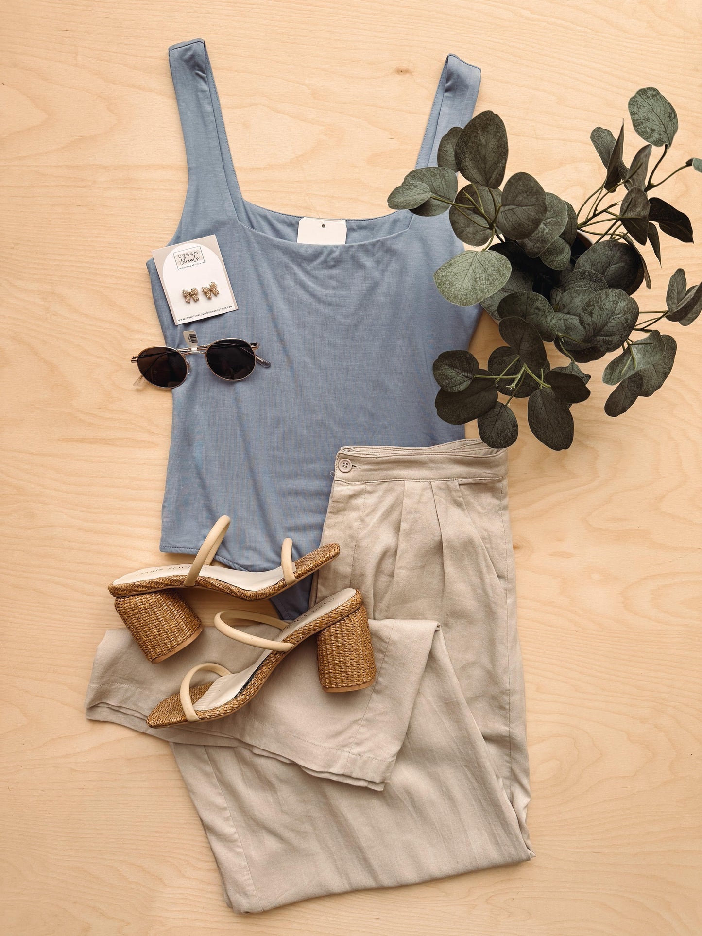 Blue tank top, beige pants, wicker sandals, sunglasses, and a plant on a wooden surface