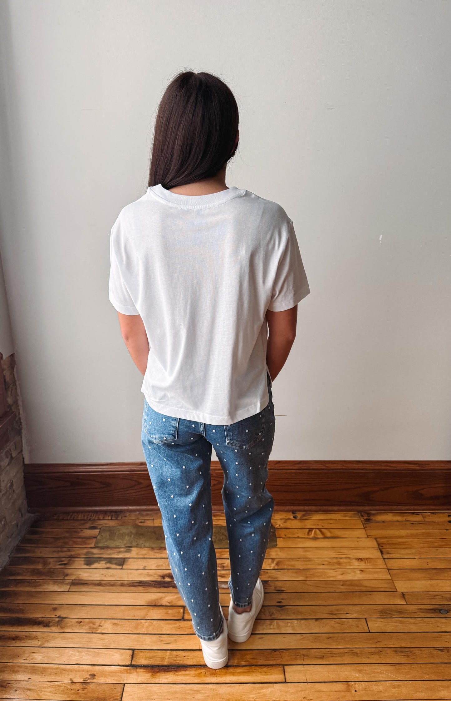 A person viewed from behind wearing a white short-sleeve t-shirt and blue jeans with small white star patterns, standing on a wooden floor against a plain wall.