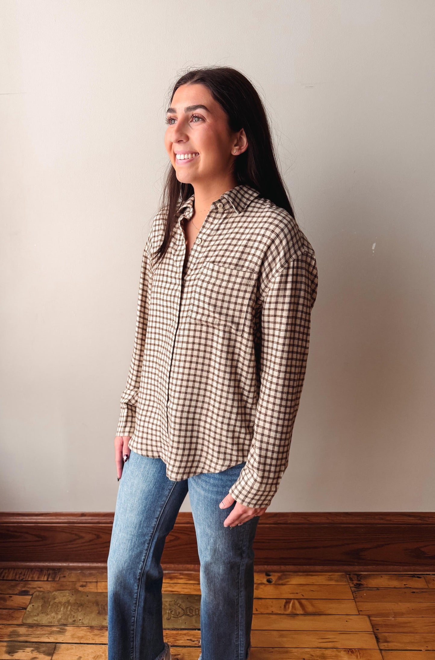 A woman wearing a brown and beige checkered button-down shirt and blue jeans, standing in a room with a beige wall and wooden floor.