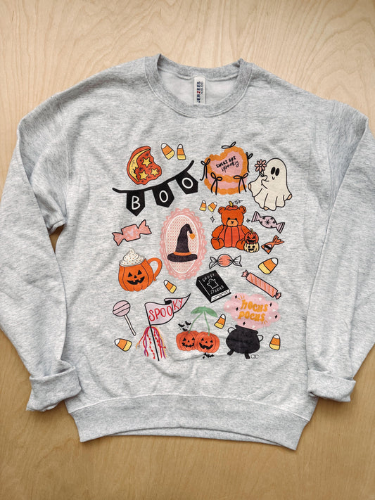 A light heather gray crewneck sweatshirt featuring a colorful Halloween-themed graphic print, laid flat on a light wood background.
