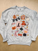 A light heather gray crewneck sweatshirt featuring a colorful Halloween-themed graphic print, laid flat on a light wood background.