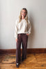 A woman stands in a room with a light gray wall and wooden floor, wearing a cream knit sweater and dark brown wide-leg drawstring pants with white side stripes.