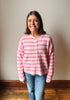 A woman smiling and wearing a pink and white striped button-up cardigan sweater with blue jeans, standing in front of a plain beige wall and wooden floor.