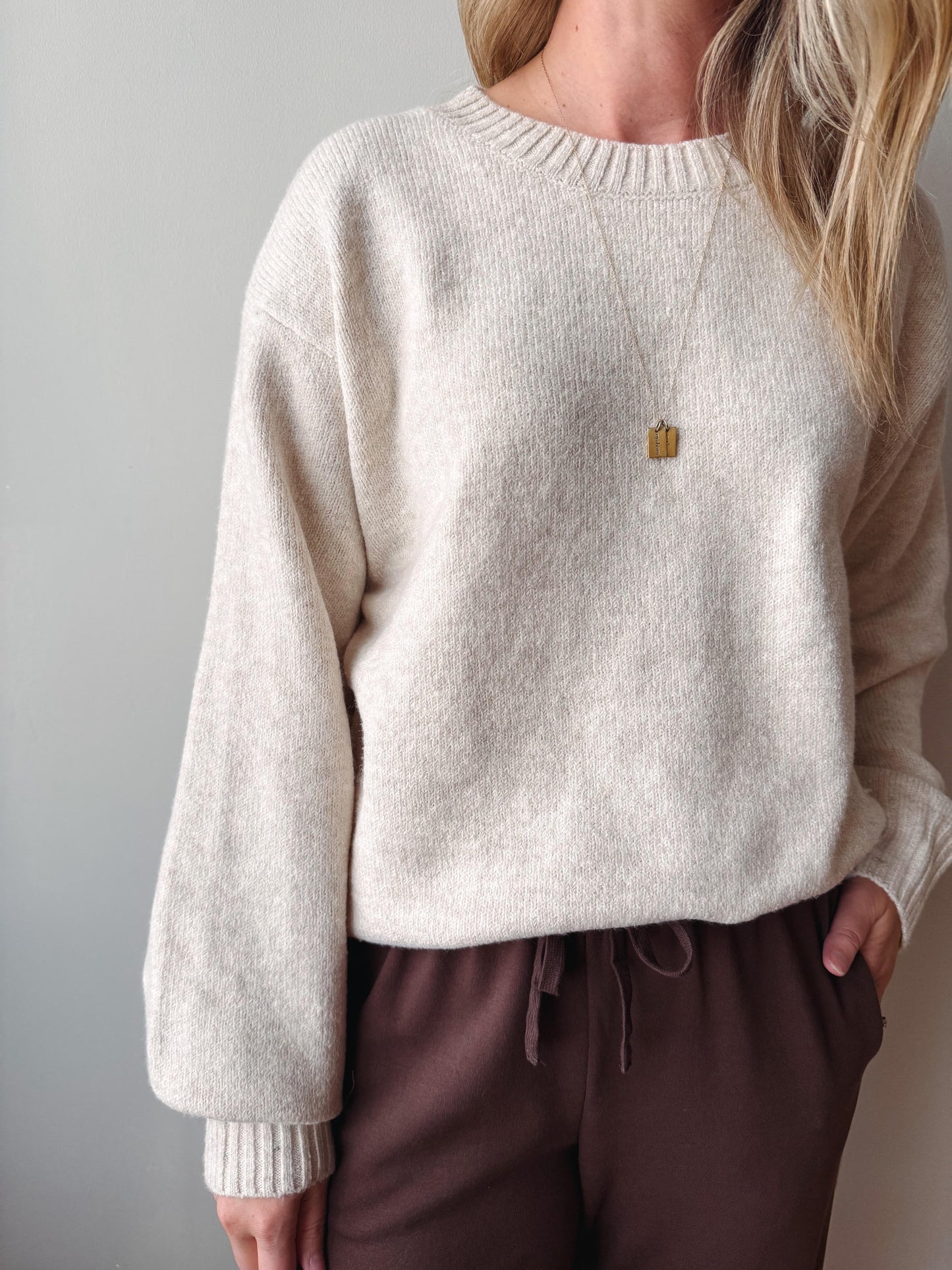 A woman wearing a cream-colored, long-sleeve knit sweater with ribbed cuffs and collar, paired with dark brown drawstring sweatpants and a delicate gold pendant necklace.