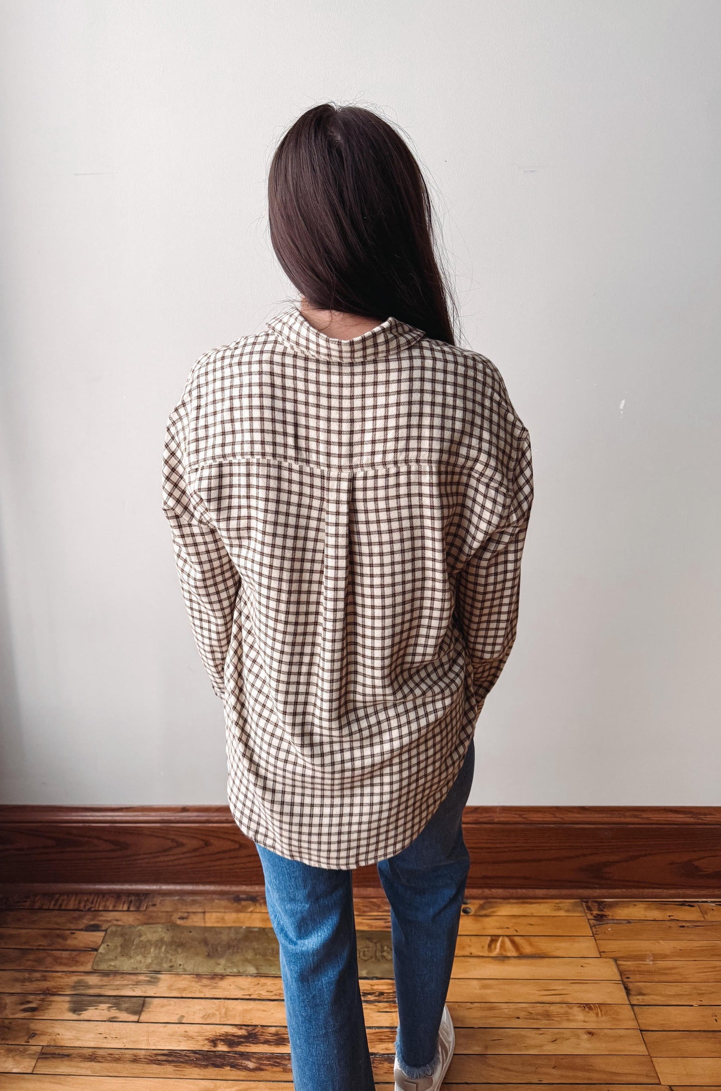 A person viewed from behind wearing an oversized brown and beige gingham plaid button-up shirt, blue jeans, and white sneakers, standing on a wooden floor against a plain white wall.