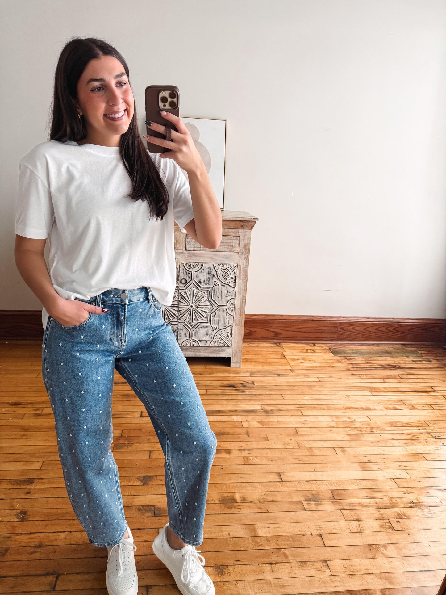 A woman taking a mirror selfie in a room with hardwood floors and a white wall. She is wearing a white short-sleeve t-shirt, light-wash blue jeans with small white polka dots, and white sneakers.