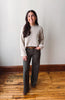 A woman stands in a room with a light gray wall and wooden floor, wearing a cream cable-knit sweater, dark gray wide-leg pants, and brown boots.