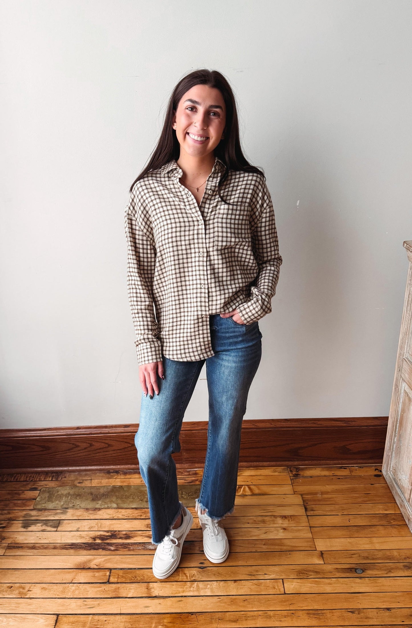 A woman stands in a room with light-colored walls and wooden floors, wearing a brown and beige gingham long-sleeve button-up shirt, blue jeans, and white sneakers.