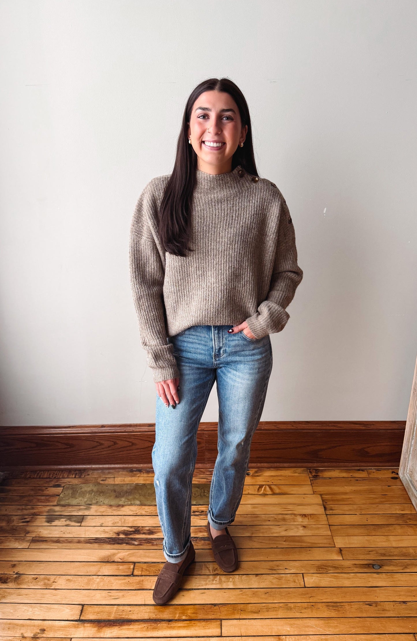 A woman stands in a room with a white wall and wooden floor, wearing a taupe ribbed knit mock neck sweater, blue jeans, and brown suede loafers.