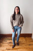 A woman stands in a room with a white wall and wooden floor, wearing a taupe ribbed knit mock neck sweater, blue jeans, and brown suede loafers.