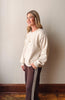 A woman wearing a cream-colored knit crewneck sweater and dark brown track pants with white side stripes, standing against a plain beige wall.
