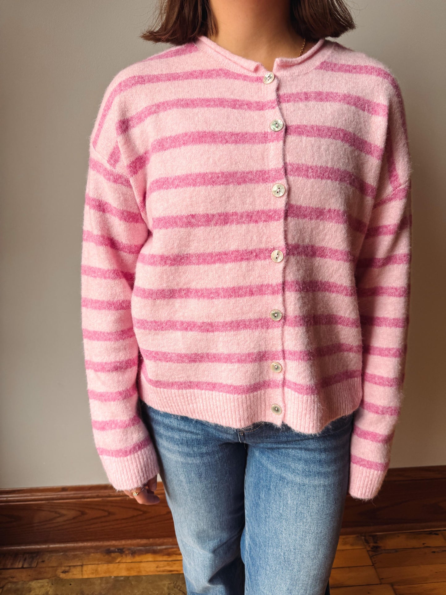 A person wearing a pink and light pink striped fuzzy knit cardigan with light-colored buttons, paired with blue jeans, standing indoors against a neutral wall.