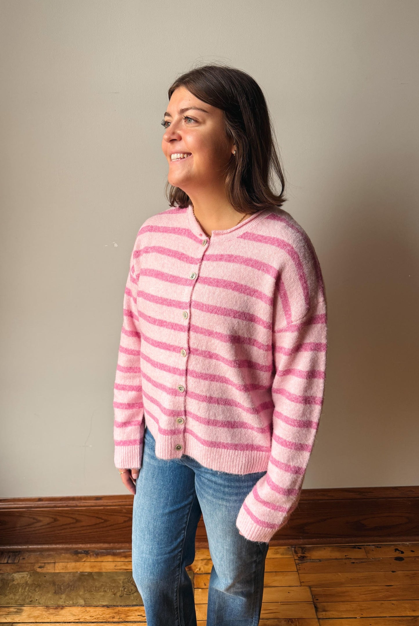 A woman wearing a pink and white striped knit cardigan sweater and blue jeans, standing in a room with a beige wall and wooden floor.