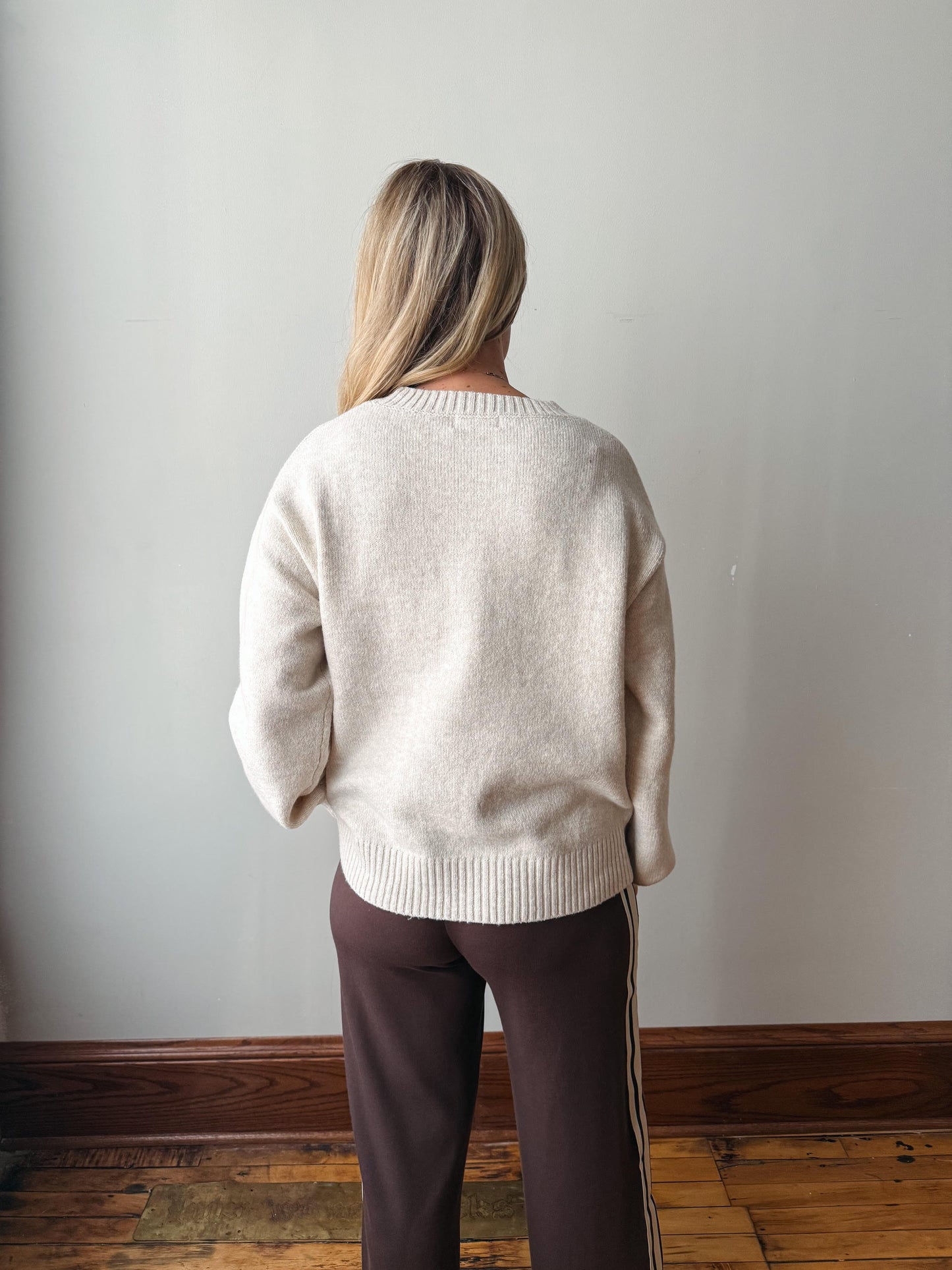 A woman viewed from behind wearing a cream-colored knit sweater and dark brown pants with a white stripe, standing against a plain light gray wall on a wooden floor.