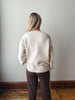 A woman viewed from behind wearing a cream-colored knit sweater and dark brown pants with a white stripe, standing against a plain light gray wall on a wooden floor.