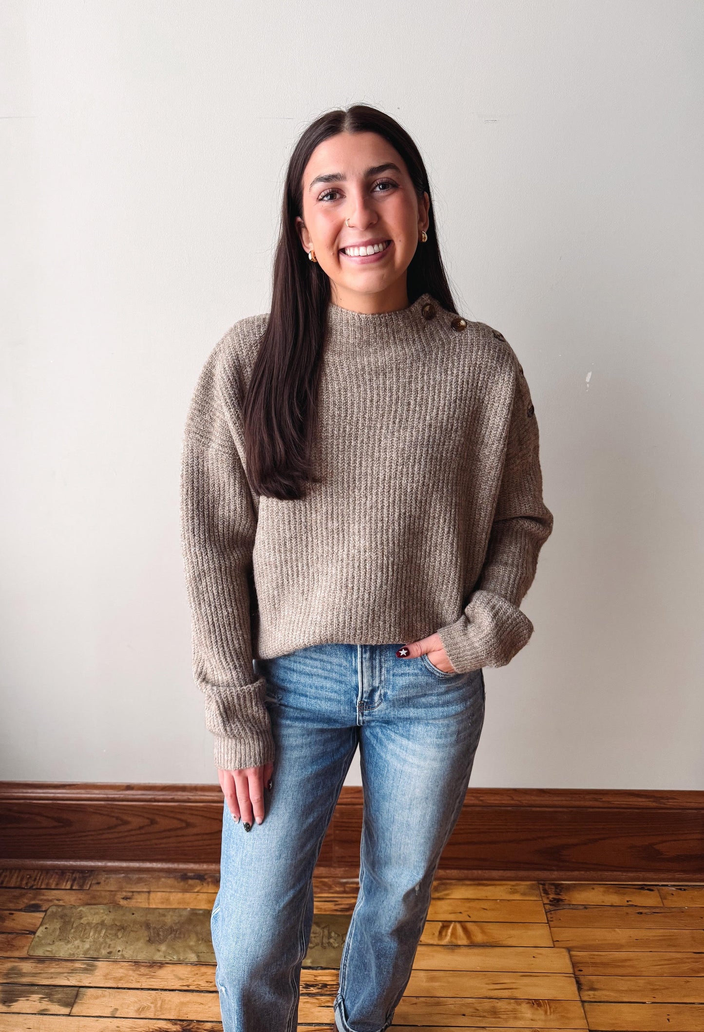 A woman wearing a taupe ribbed knit mock neck sweater and blue jeans, standing in front of a plain off-white wall with a wooden baseboard and hardwood floor.