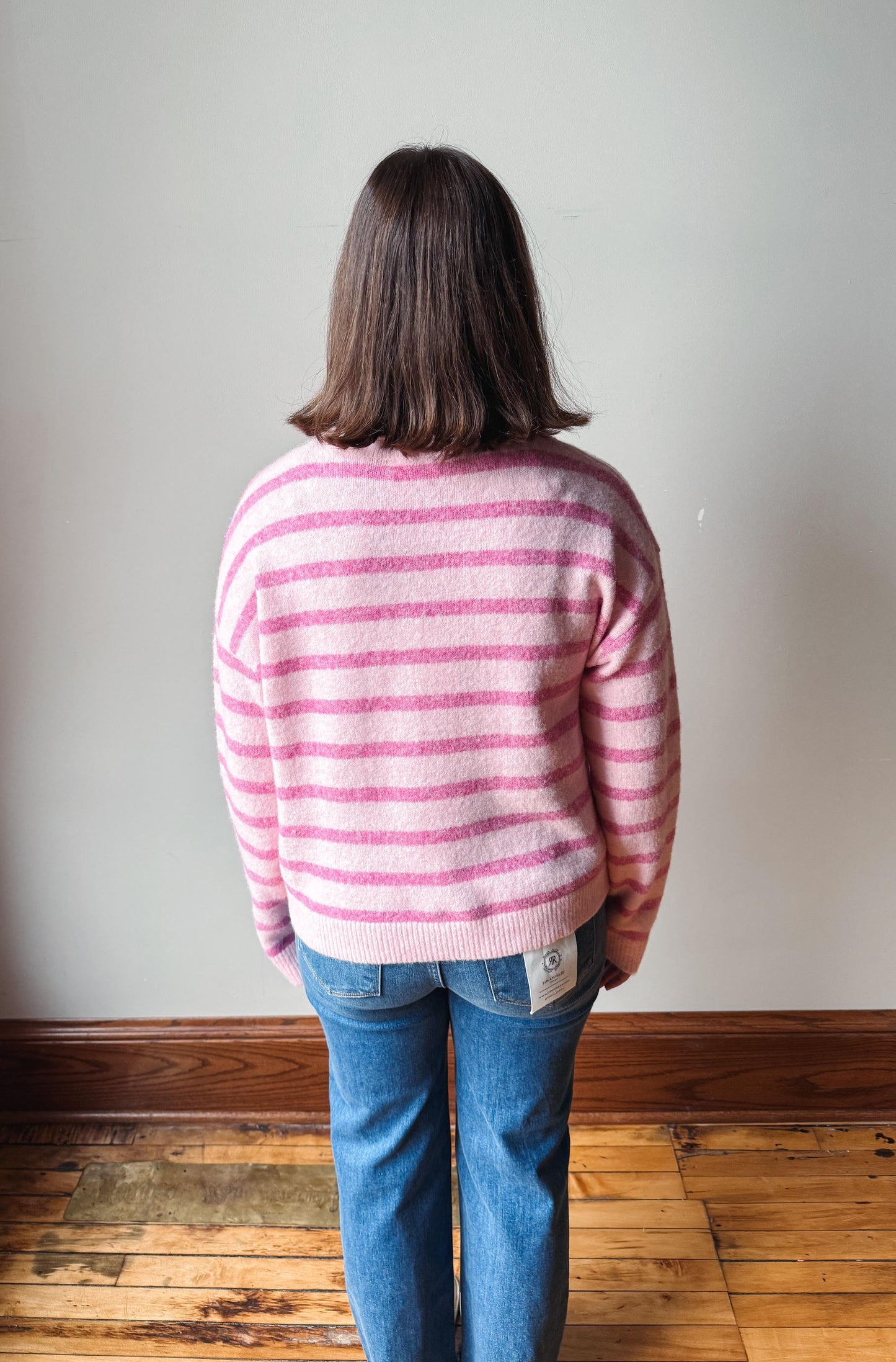 A person viewed from behind wearing a pink and white striped fuzzy knit sweater, blue jeans, and standing on a wooden floor against a plain wall.