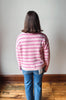 A person viewed from behind wearing a pink and white striped fuzzy knit sweater, blue jeans, and standing on a wooden floor against a plain wall.