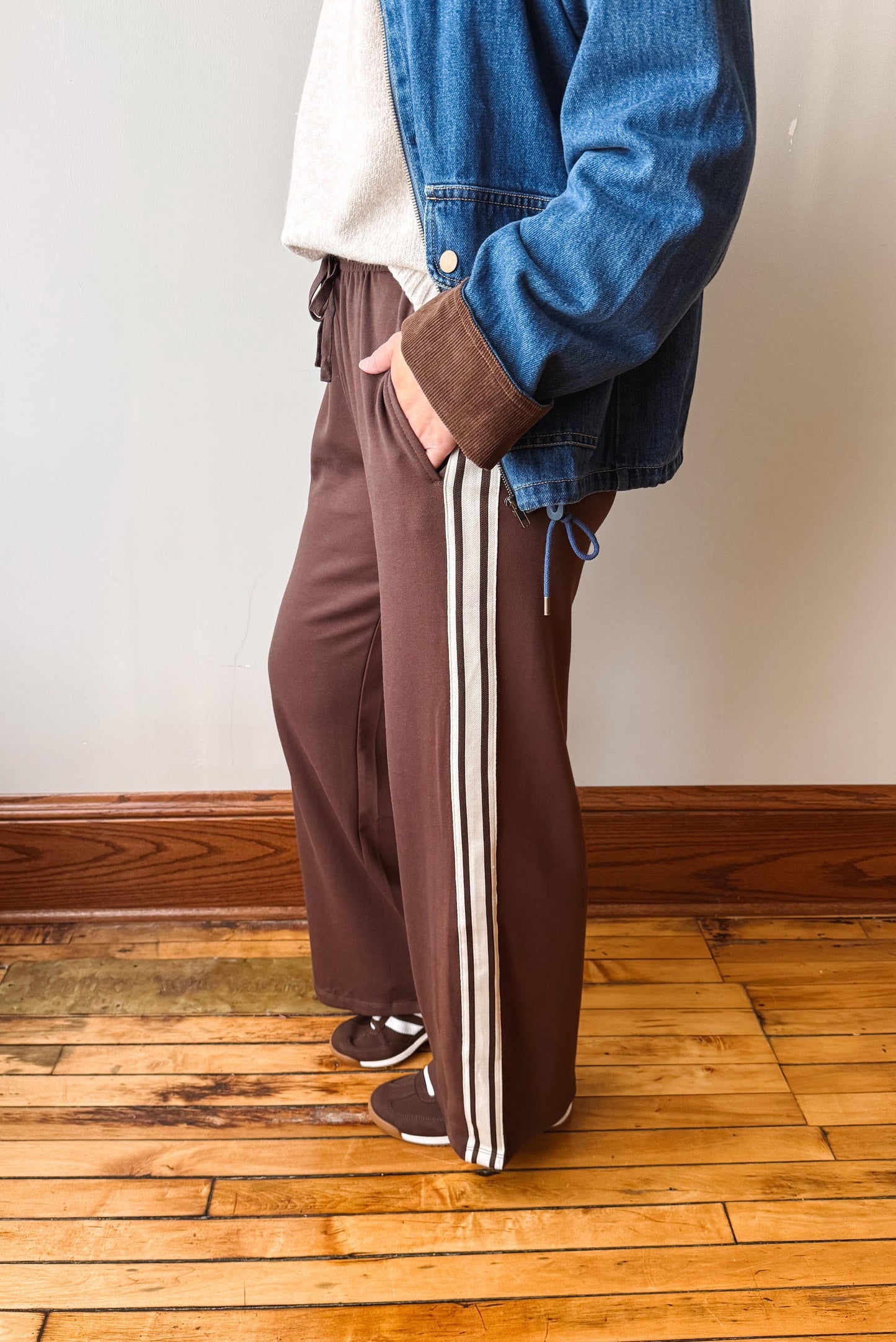 A person wearing brown wide-leg track pants with three white side stripes, styled with a blue denim jacket, cream shirt, and sneakers, standing on a wooden floor against a plain wall.