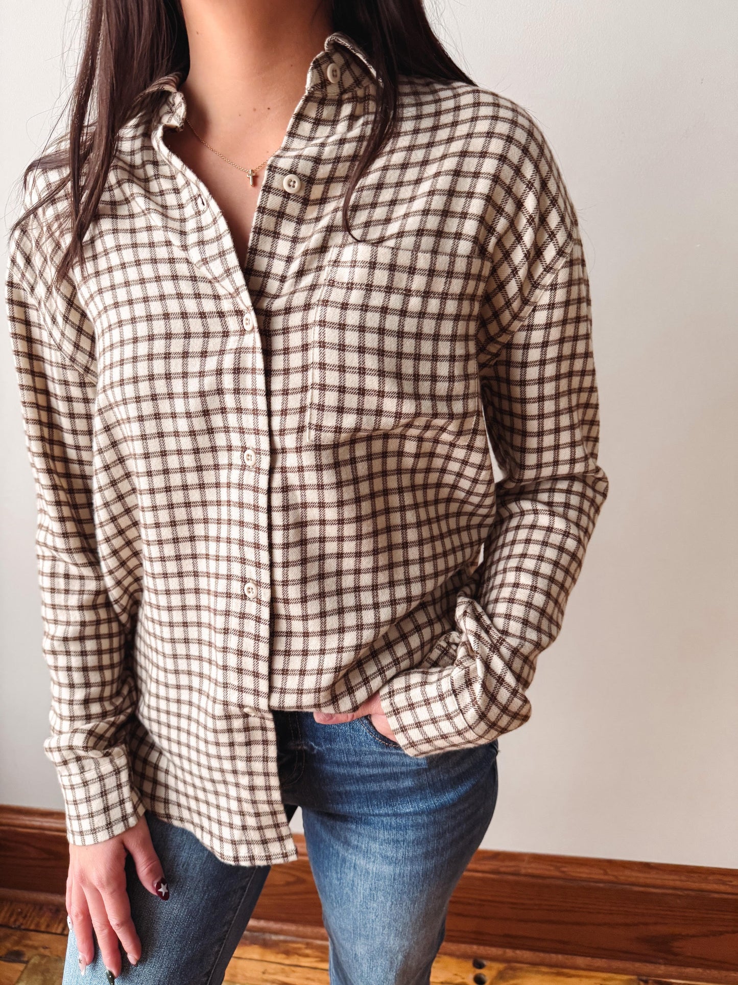 A woman wearing a brown and beige checkered long-sleeve button-up shirt with blue jeans, standing against a plain off-white wall.