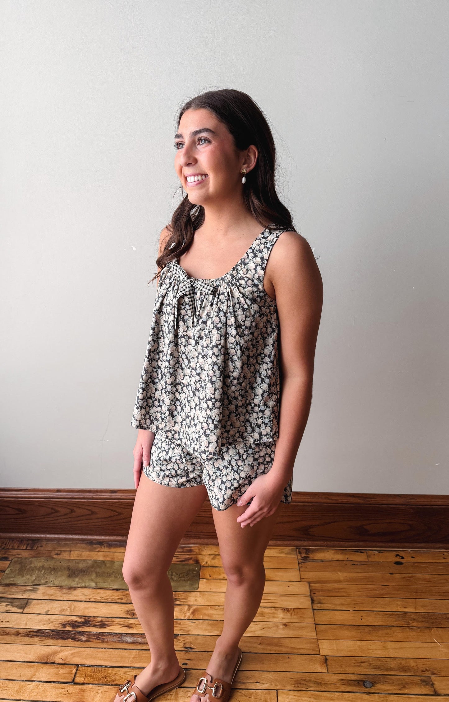 Woman wearing a floral sleeveless top and shorts standing on a wooden floor against a white wall.