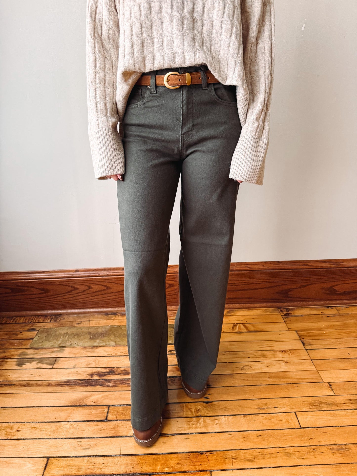 A person wearing dark charcoal gray wide-leg pants with a brown leather belt, paired with a cream cable-knit sweater and brown shoes, standing on a wooden floor against a white wall.
