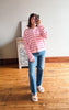 A woman takes a mirror selfie wearing a pink and white striped zip-up cardigan, blue jeans, and white slide sandals in a room with a wooden floor and a light-colored wall.
