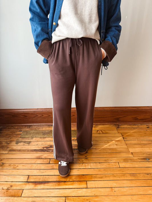 A person wearing brown wide-leg sweatpants with a drawstring waist, styled with a cream sweater, a blue jacket, and brown slip-on shoes, standing on a wooden floor against a plain wall.