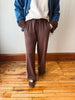 A person wearing brown wide-leg sweatpants with a drawstring waist, styled with a cream sweater, a blue jacket, and brown slip-on shoes, standing on a wooden floor against a plain wall.