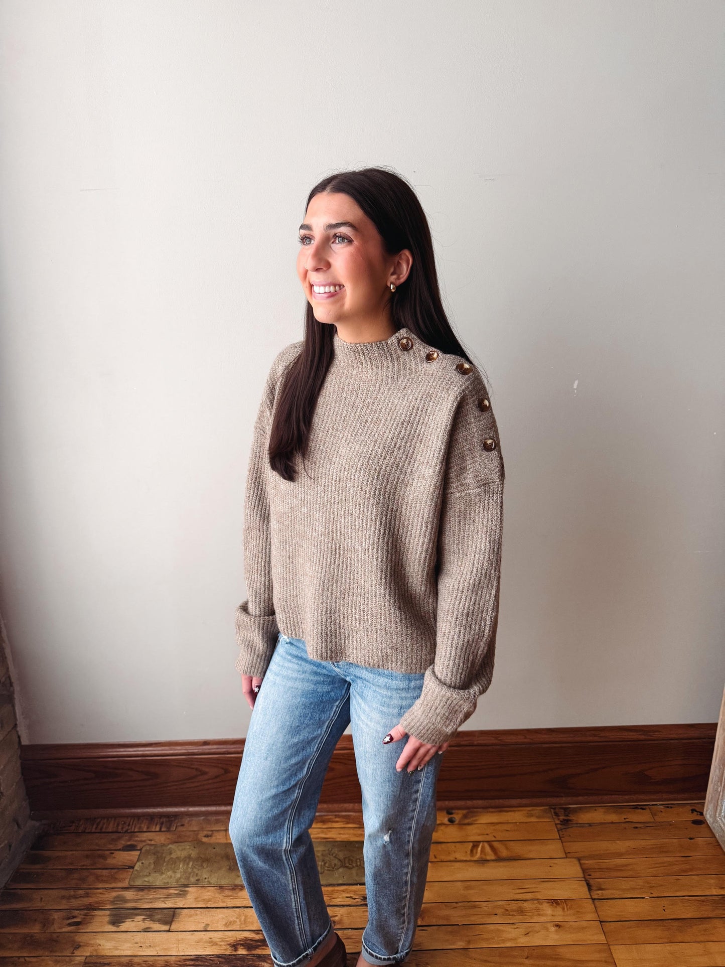 A woman wearing a beige ribbed knit mock neck sweater with decorative buttons on the shoulders and light blue jeans, standing in a room with a light gray wall and wooden floor.