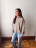 A woman wearing a beige ribbed knit mock neck sweater with decorative buttons on the shoulders and light blue jeans, standing in a room with a light gray wall and wooden floor.