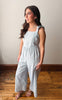 Woman wearing a light blue jumpsuit standing on a wooden floor with a plain background