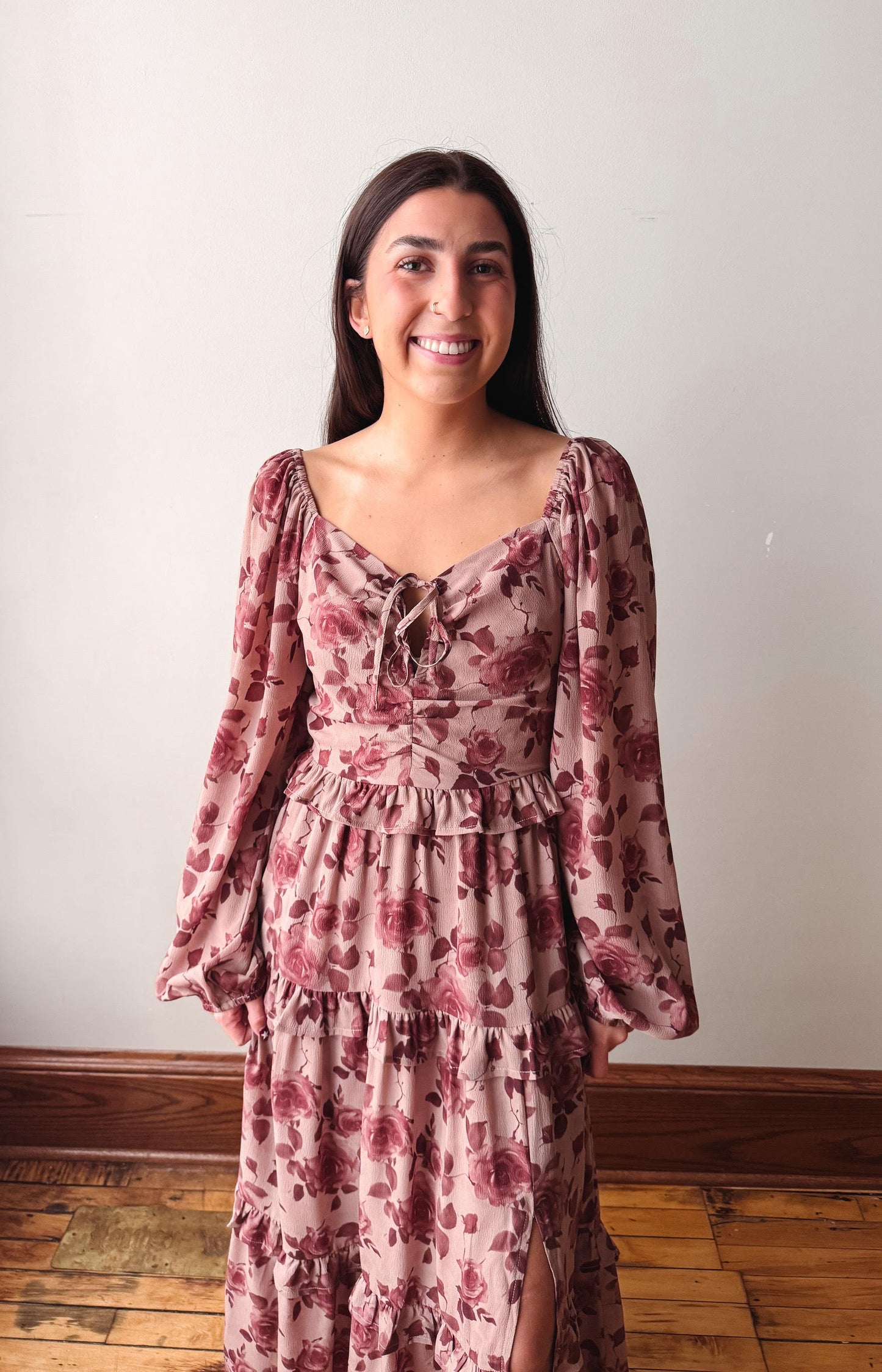 A woman smiling and wearing a dusty rose floral long-sleeve maxi dress with a tiered skirt, standing in front of a plain off-white wall and wooden floor.