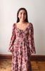 A woman smiling and wearing a dusty rose floral long-sleeve maxi dress with a tiered skirt, standing in front of a plain off-white wall and wooden floor.