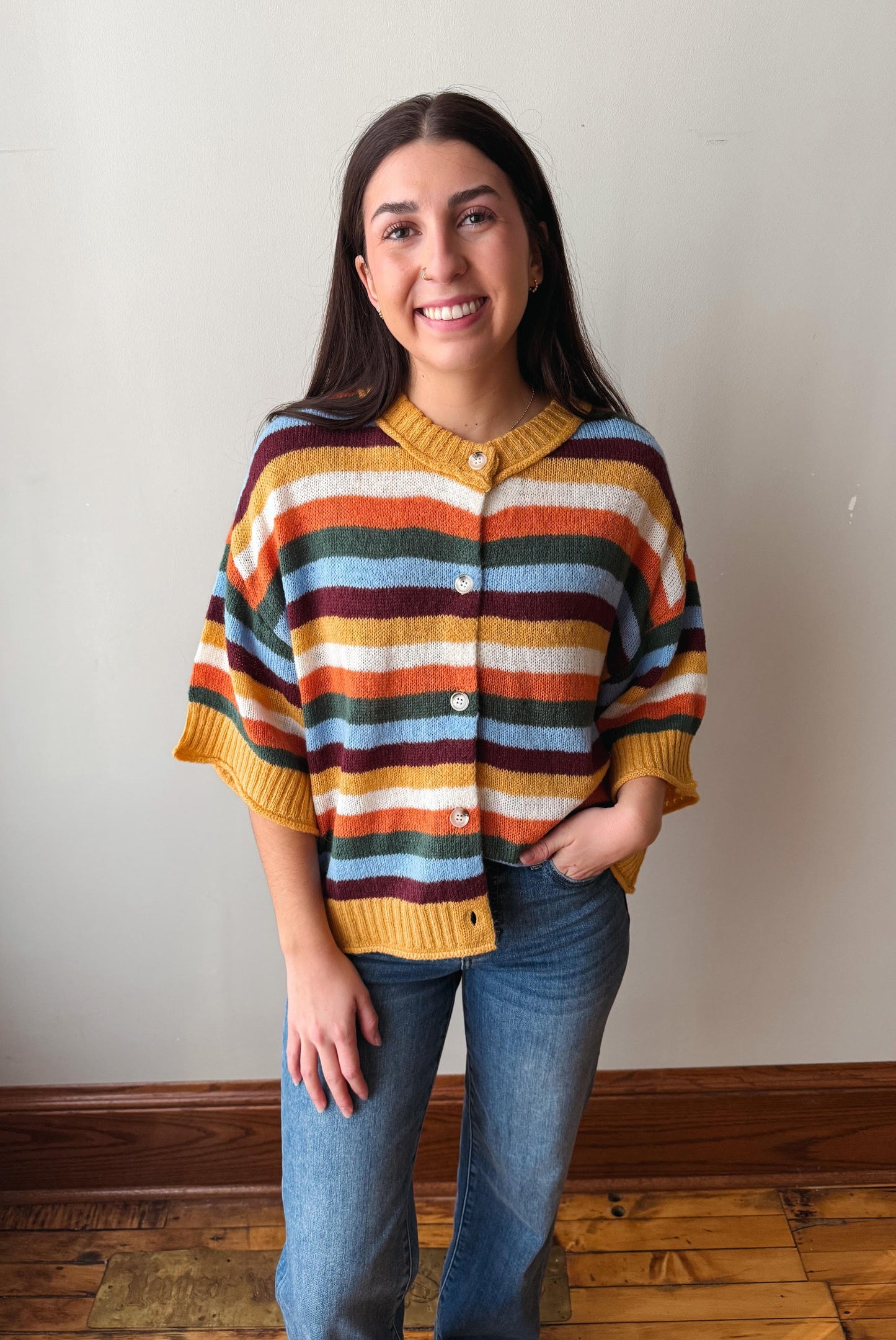 A woman wearing a colorful striped knit cardigan sweater with blue jeans, standing against a plain off-white wall with a wooden floor visible.